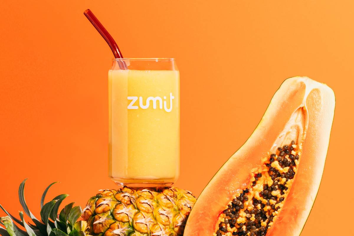 Smoothie Zumit Caribbean Passion 100% natural Smoothie Zumit Caribbean Passion con mango, papaya y piña apoyado sobre una piña y una papaya